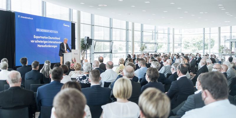 männlichen Moderator steht auf einer Bühne, davor sitzt ein Publikum 